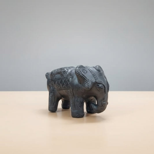 A small carved elephant figurine in Sawai Madhopur Black Pottery crafts from Rajasthan, India, standing on a woven beige and white placemat against a plain light gray background.